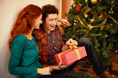 Buy stock photo Shot of a young man opening his christmas present while sitting with his girlfriend