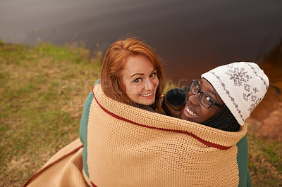 Buy stock photo A high angle portrait of two happy young women huddled under a blanket next to a lake in winter