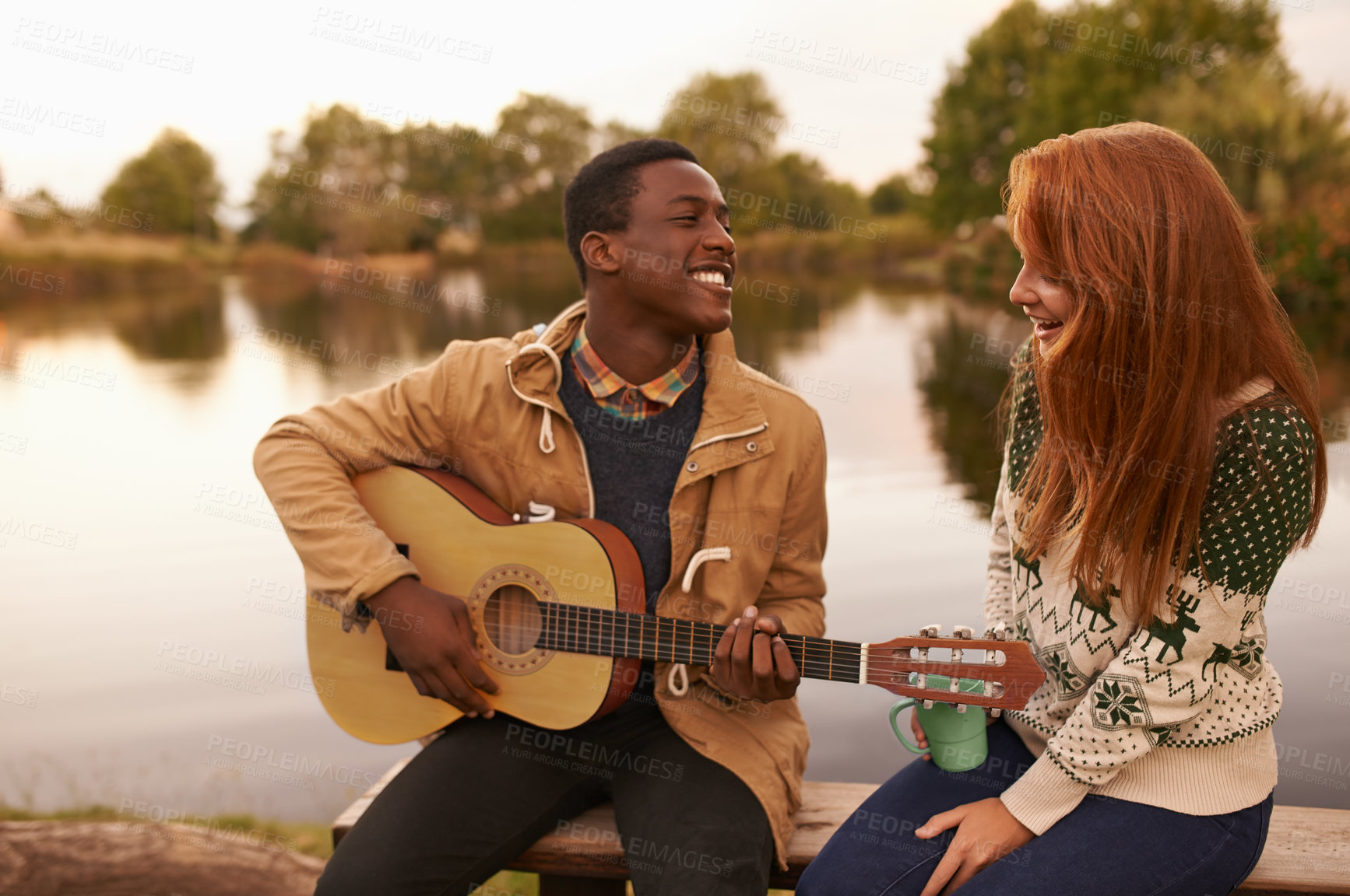 Buy stock photo A handsome young man serenading a beautiful girl while sitting next to a lake