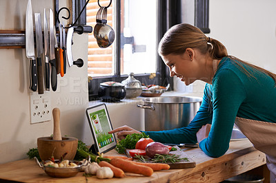Buy stock photo Shot of a young woman looking at an online recipe on her digital tablet while preparing a meal
