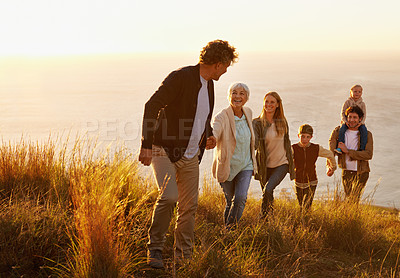 Buy stock photo A multi-generational family walking up a grassy hill together at sunset with the ocean in the background