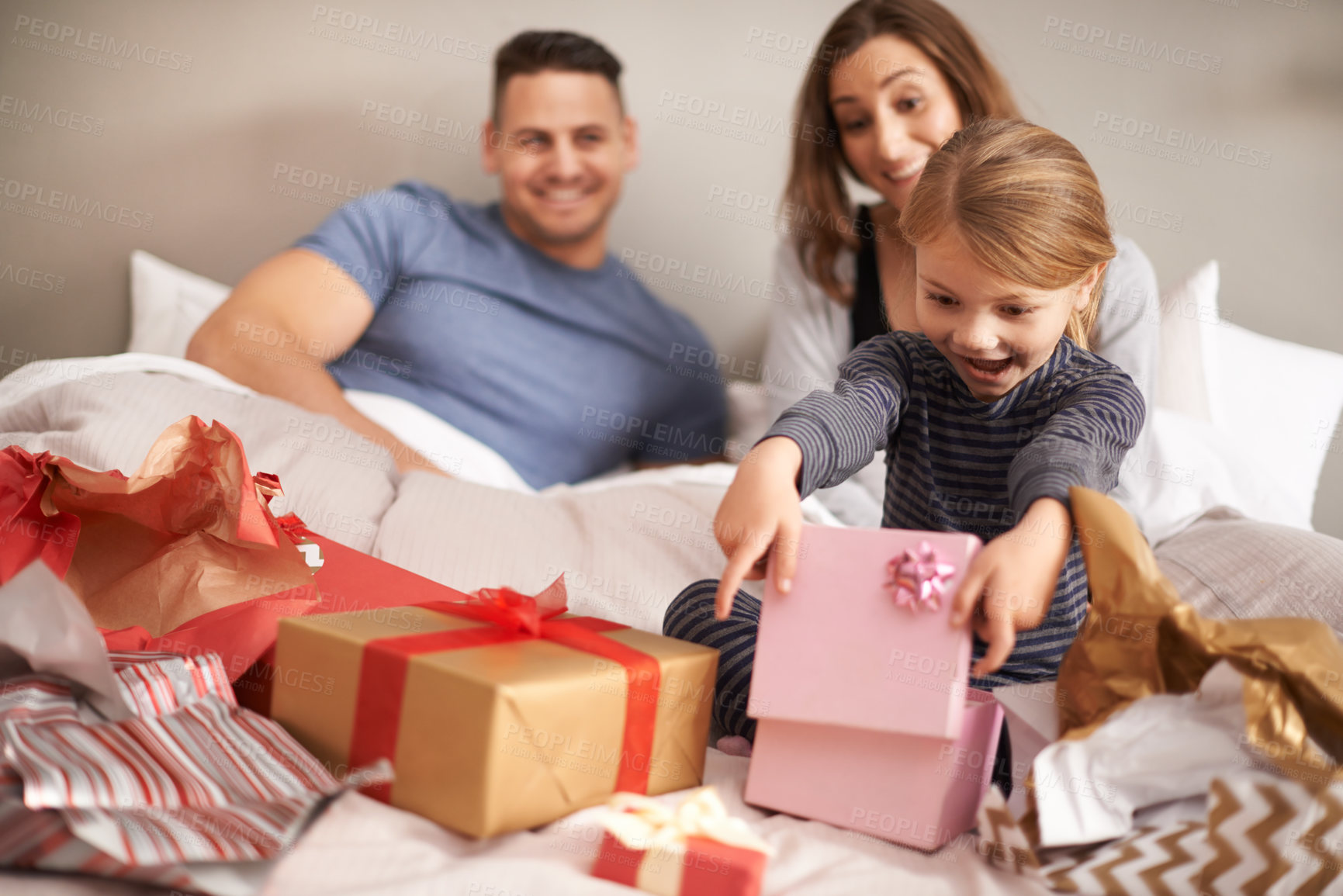 Buy stock photo Shot of a little girl opening presents in bed as her parents watch
