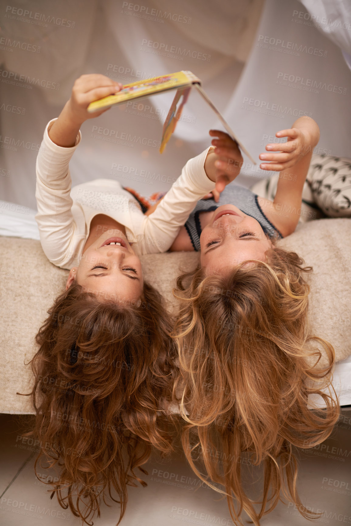 Buy stock photo Shot of two little girls reading a book while lying upside down on a bed