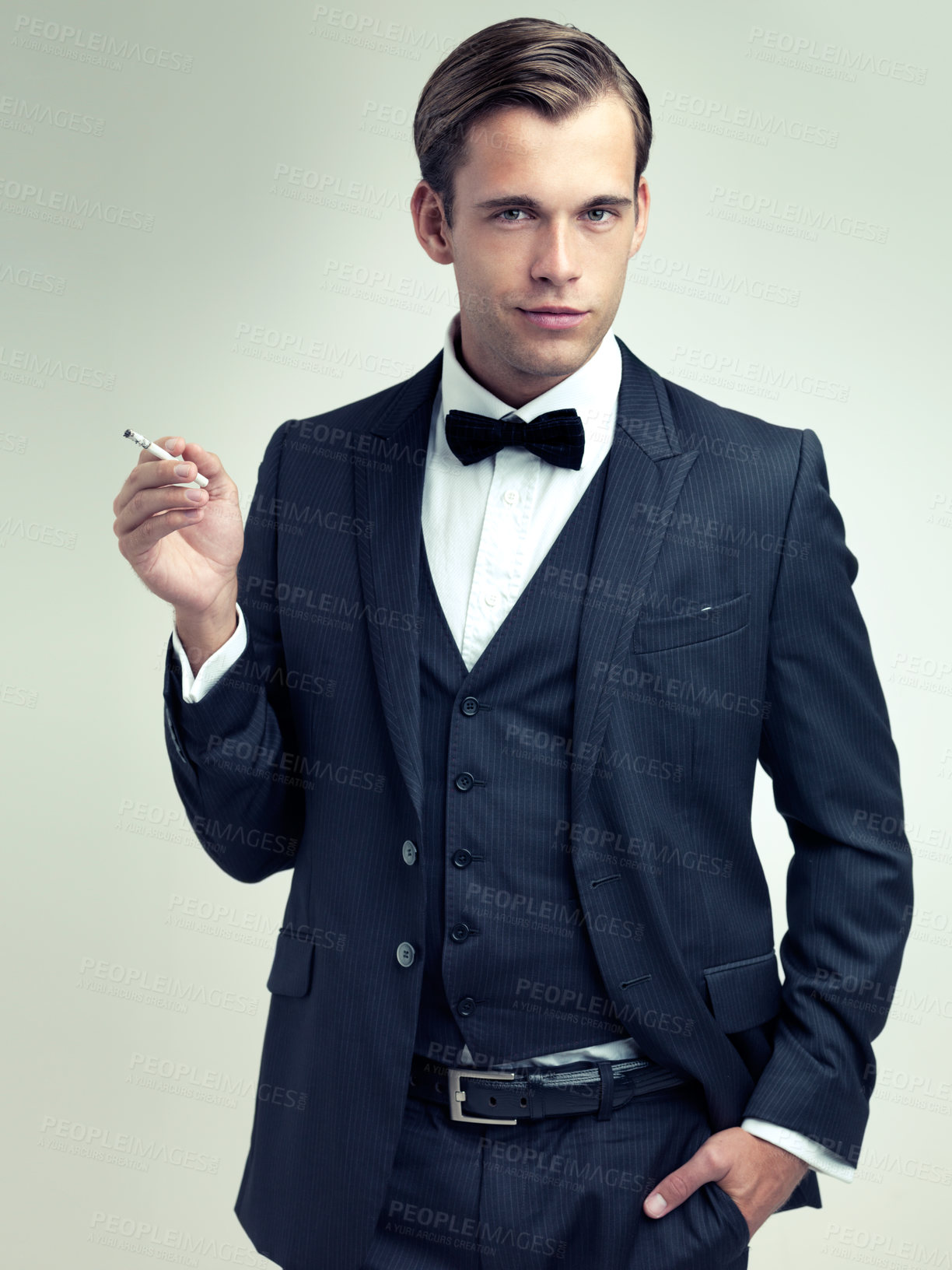 Buy stock photo Portrait, elegant or young gentleman smoking a cigarette in studio, confidence or vintage fashion by white background. Face, serious and man in retro suit or tuxedo, bow tie and class at formal event