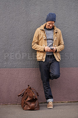Buy stock photo Shot of a handsome young man sending a text message while leaning against a wall outside