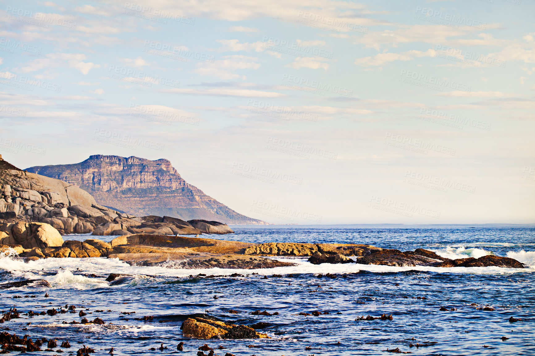 Buy stock photo Mountains, ocean and nature with waves at beach for environment, landscape and blue sky. Calm, summer and seascape with sunrise on horizon at coastline for tropical, clouds and water on rock surface