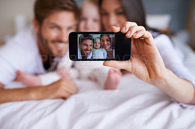 Buy stock photo Shot of a young couple lying on the bed with their baby girl and taking a selfie with their mobile phone