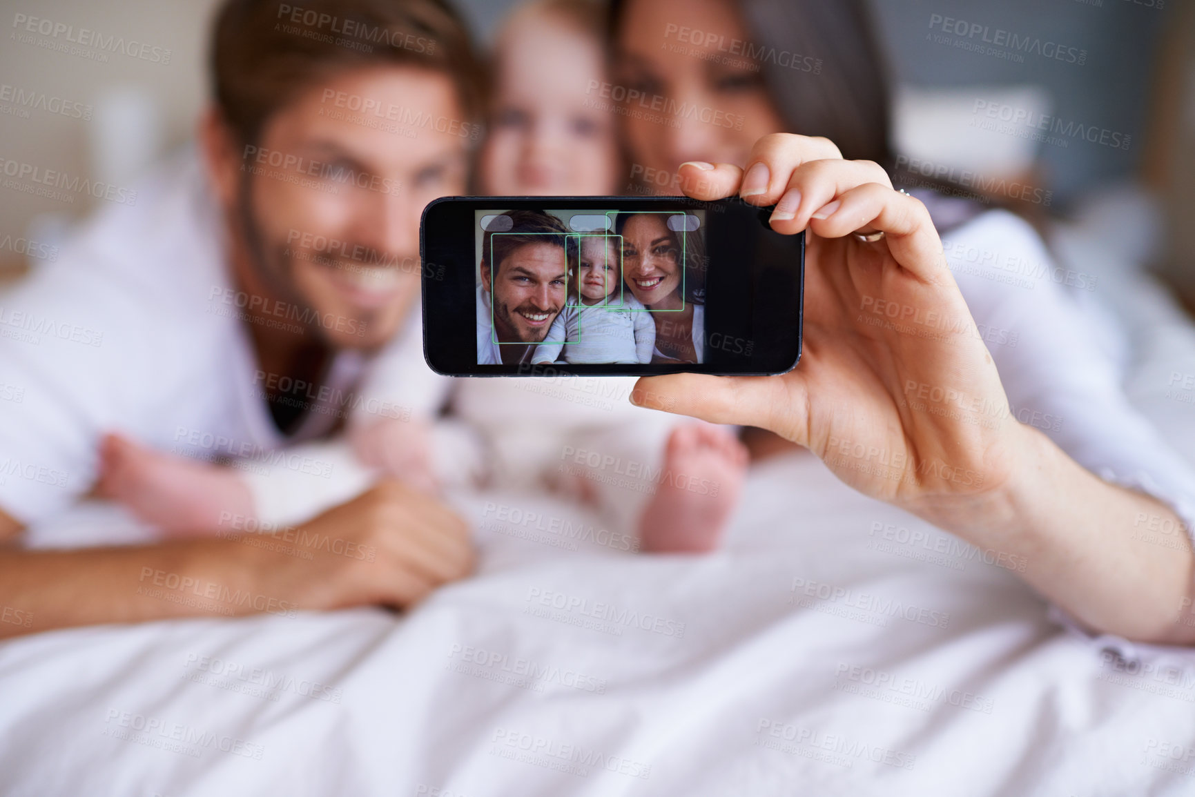 Buy stock photo Shot of a young couple lying on the bed with their baby girl and taking a selfie with their mobile phone