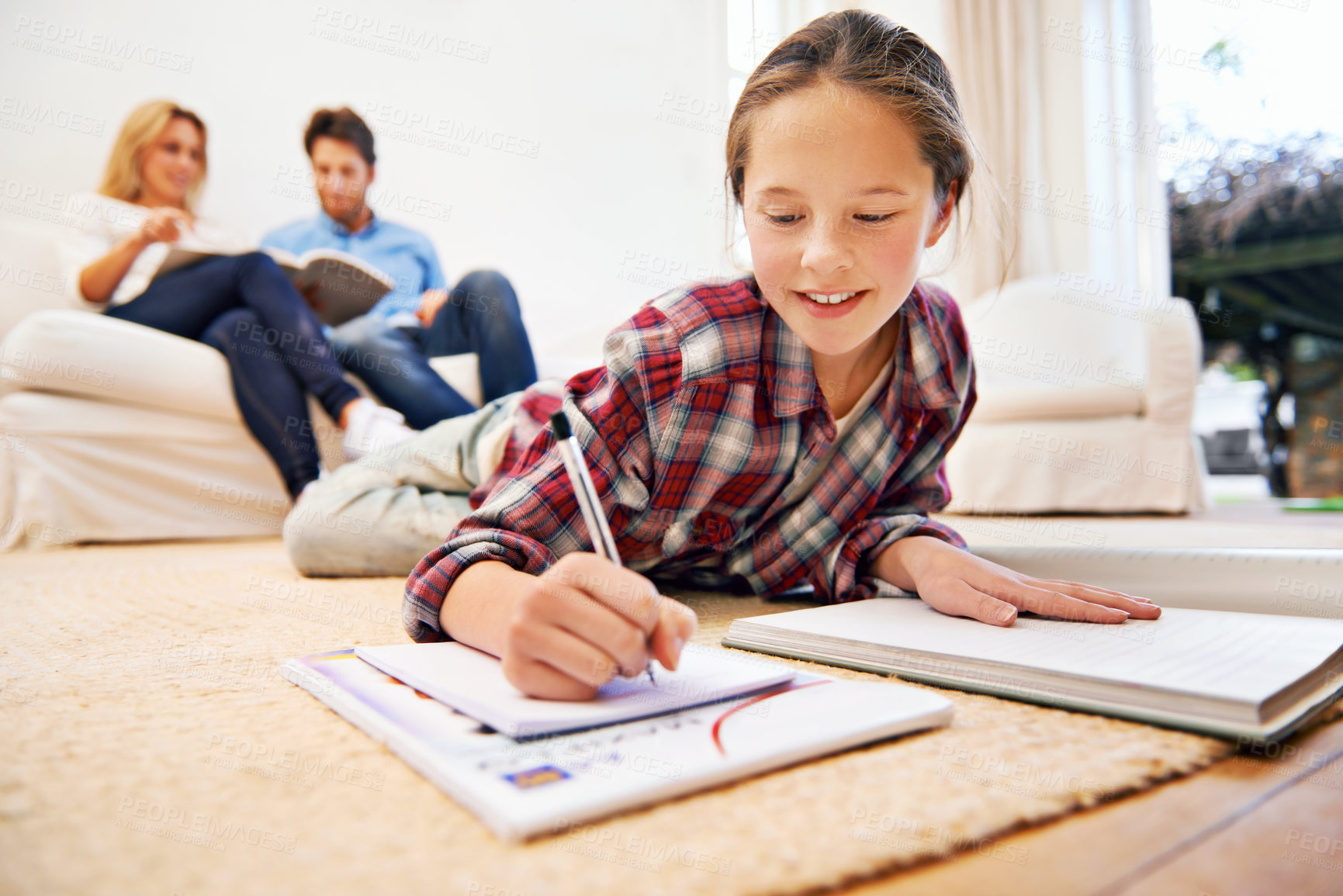 Buy stock photo Shot of a little girl lying on the living room floor doing homework with her parents in the background
