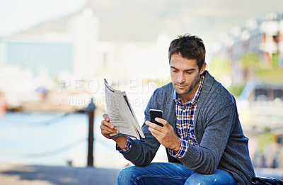 Buy stock photo Shot of a handsome man holding the newspaper and using his phone outdoors