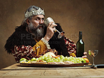 Buy stock photo King, confident and feast at table with wine glass as royalty in studio on brown background for tradition, culture and meal. Portrait, monarch and leader with eating buffet or supper with confidence
