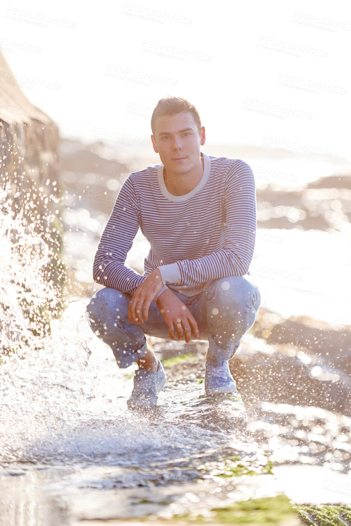 Buy stock photo Ocean, water and portrait of male man, sunset and nature for vacation or holiday. Confidence, splash and wave from beach on rocky terrain while travel, seaside and sunlight on coastline getaway