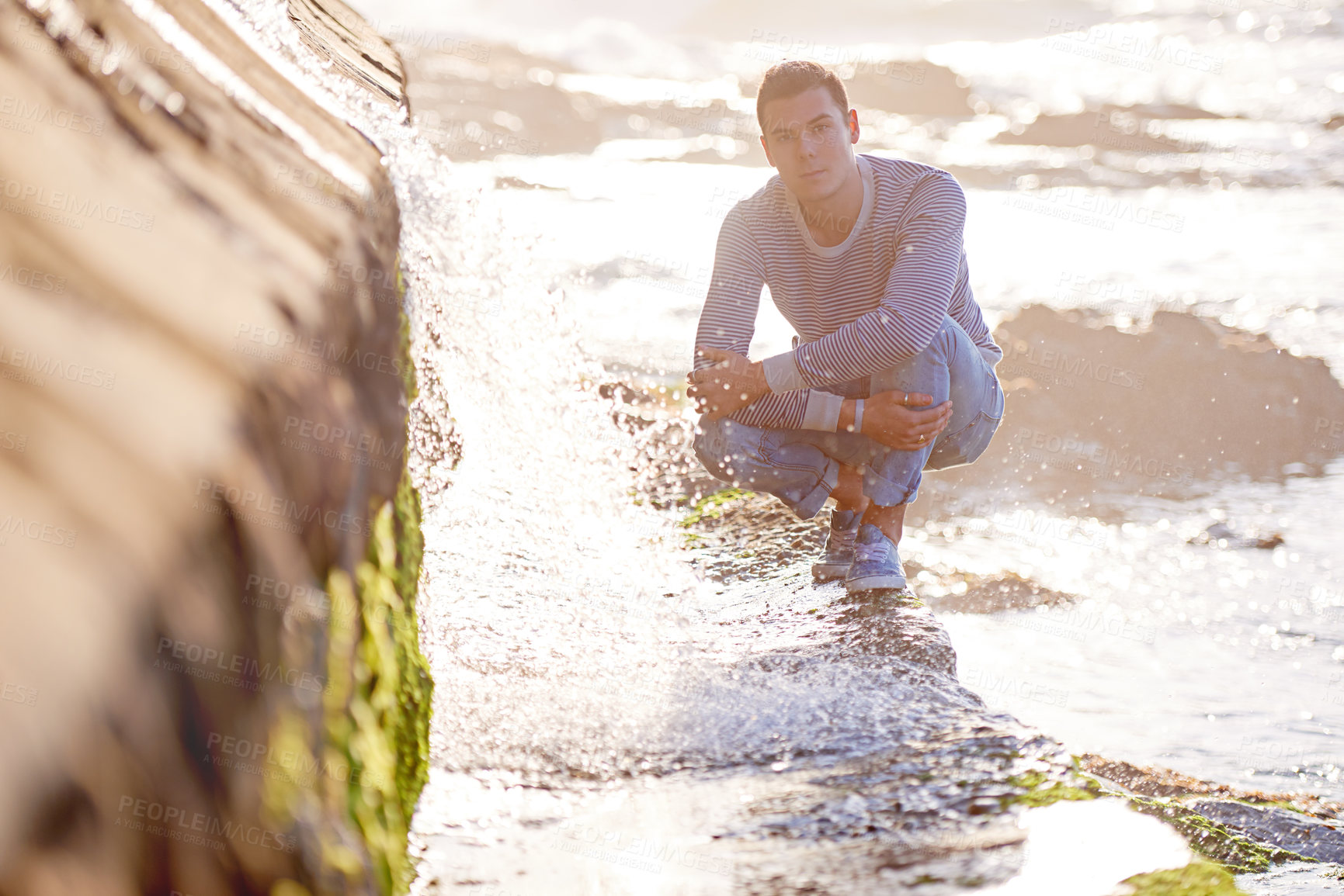 Buy stock photo Handsome man, portrait and beach with water in relax for summer holiday, weekend or enjoying day by the sea. Young male person crouching on rocky slope by ocean waves for outdoor vacation in nature