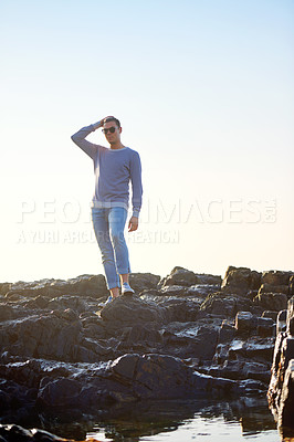 Buy stock photo Man, rocks and water for fashion with summer vacation, confidence and sunglasses at beach. Male person, holiday and traveling at seaside for sunlight, relaxation and trendy clothes with eyewear