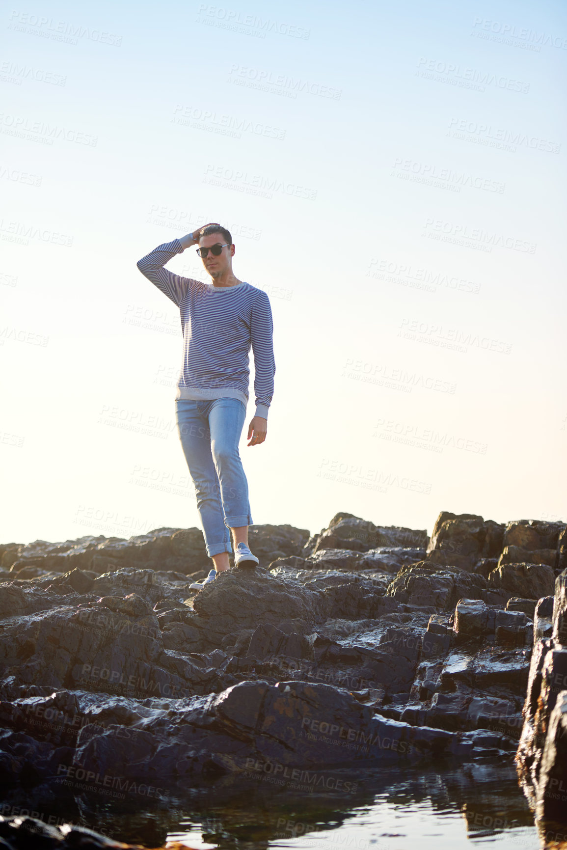 Buy stock photo Man, rocks and water for fashion with summer vacation, confidence and sunglasses at beach. Male person, holiday and traveling at seaside for sunlight, relaxation and trendy clothes with eyewear