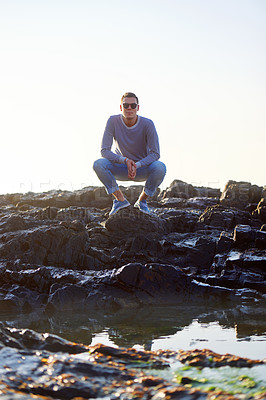 Buy stock photo Man, rocks and water for fashion at beach with summer holiday, trendy and sunglasses. Male tourist, stylish and travel at seaside for sunlight, relaxation and confidence with eyewear in Cape Town 