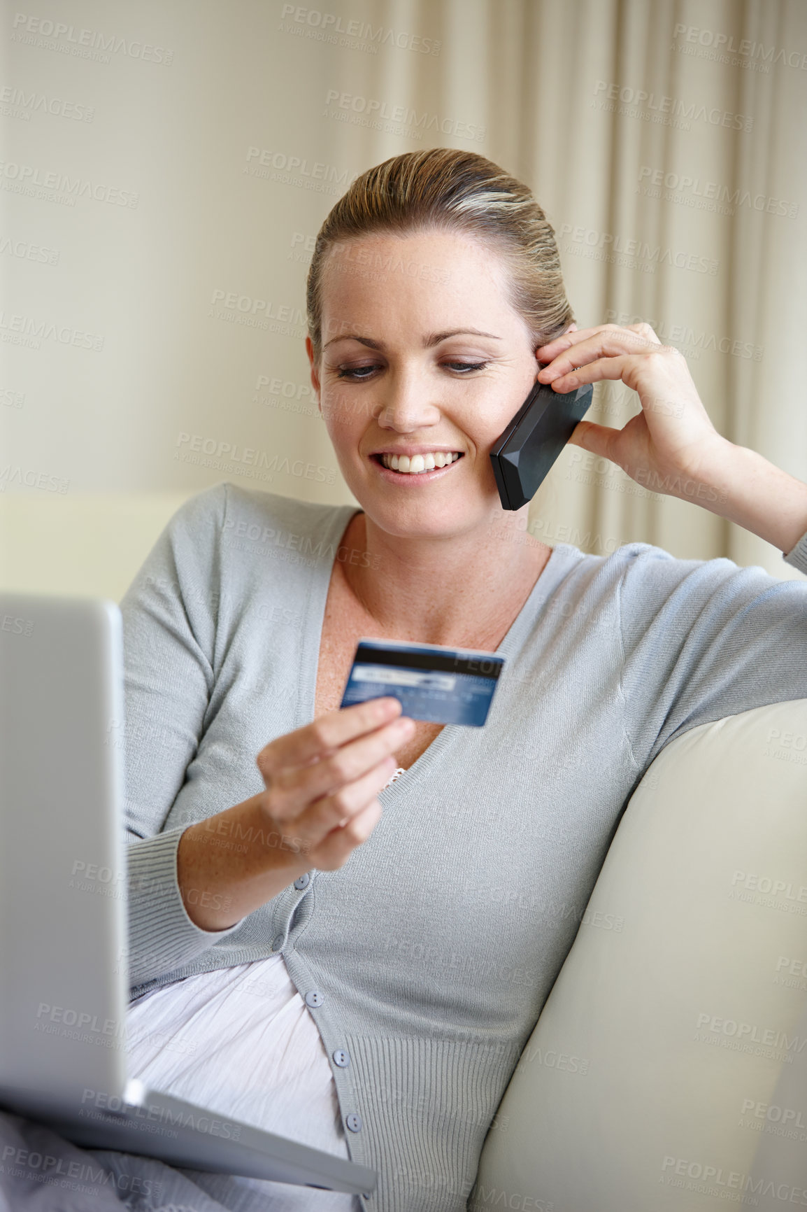 Buy stock photo A young woman talking on her cellphone while holding a bank card and sitting with a laptop