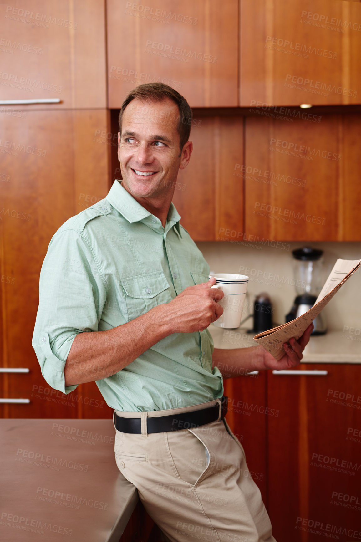 Buy stock photo A happy man holding a newspaper and a mug while leaning against a counter top in a kitchen