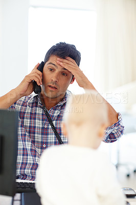 Buy stock photo Shot of a stressed out working dad on the phone with his baby sitting in front of him 
