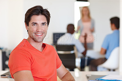 Buy stock photo A trendy businessman smiling while looking at the camera while sitting in an office with his colleagues in the background. One confident casually dressed male entrepreneur working in a creative office