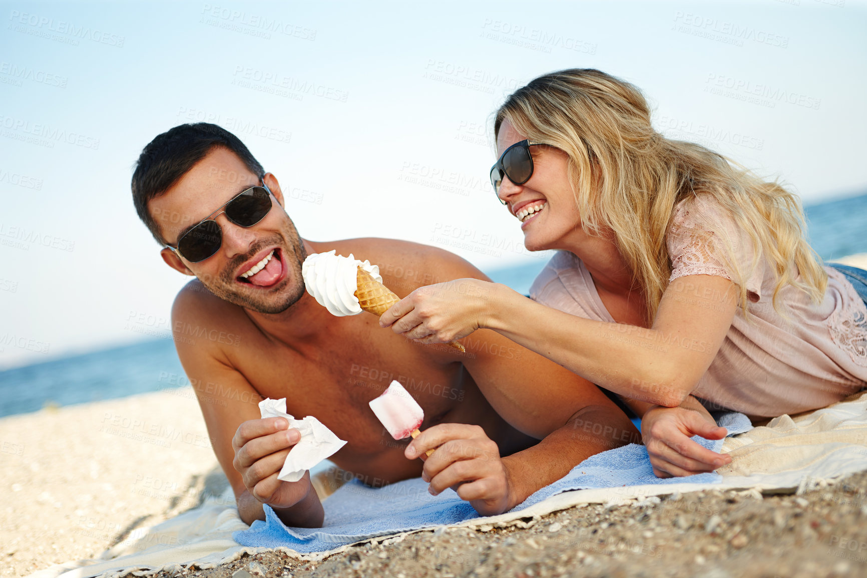Buy stock photo Shot of a happy young couple eating ice cream while lying on a sunny beach