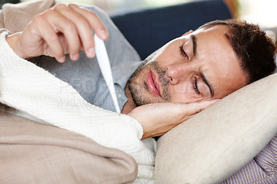 Buy stock photo Man suffering cold and flu symptoms, worried about corona virus and stressed about taking leave from work to recover. A young man lying on the sofa reading his temperature on a thermometer. 