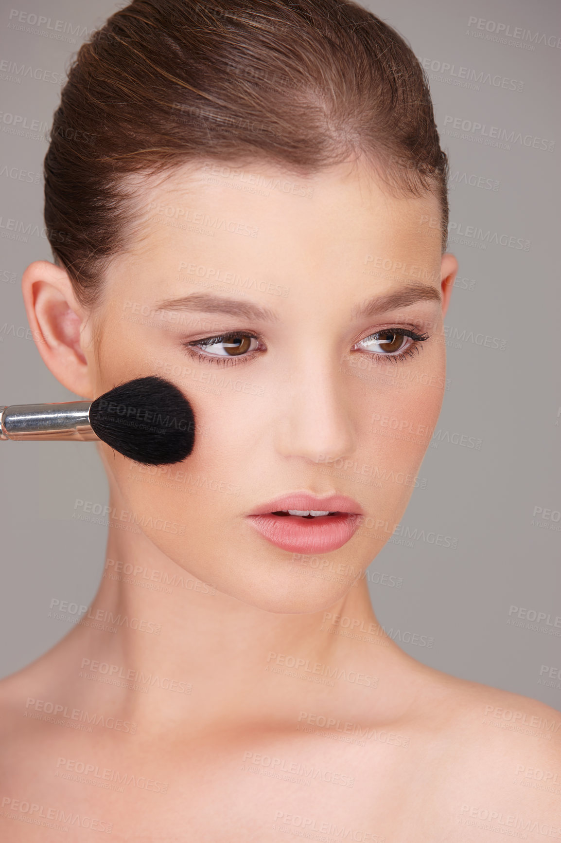 Buy stock photo A beautiful woman applying blush to her cheeks with a round makeup brush. A gorgeous female applying makeup while grooming and doing a skin care routine in a studio against grey copyspace background