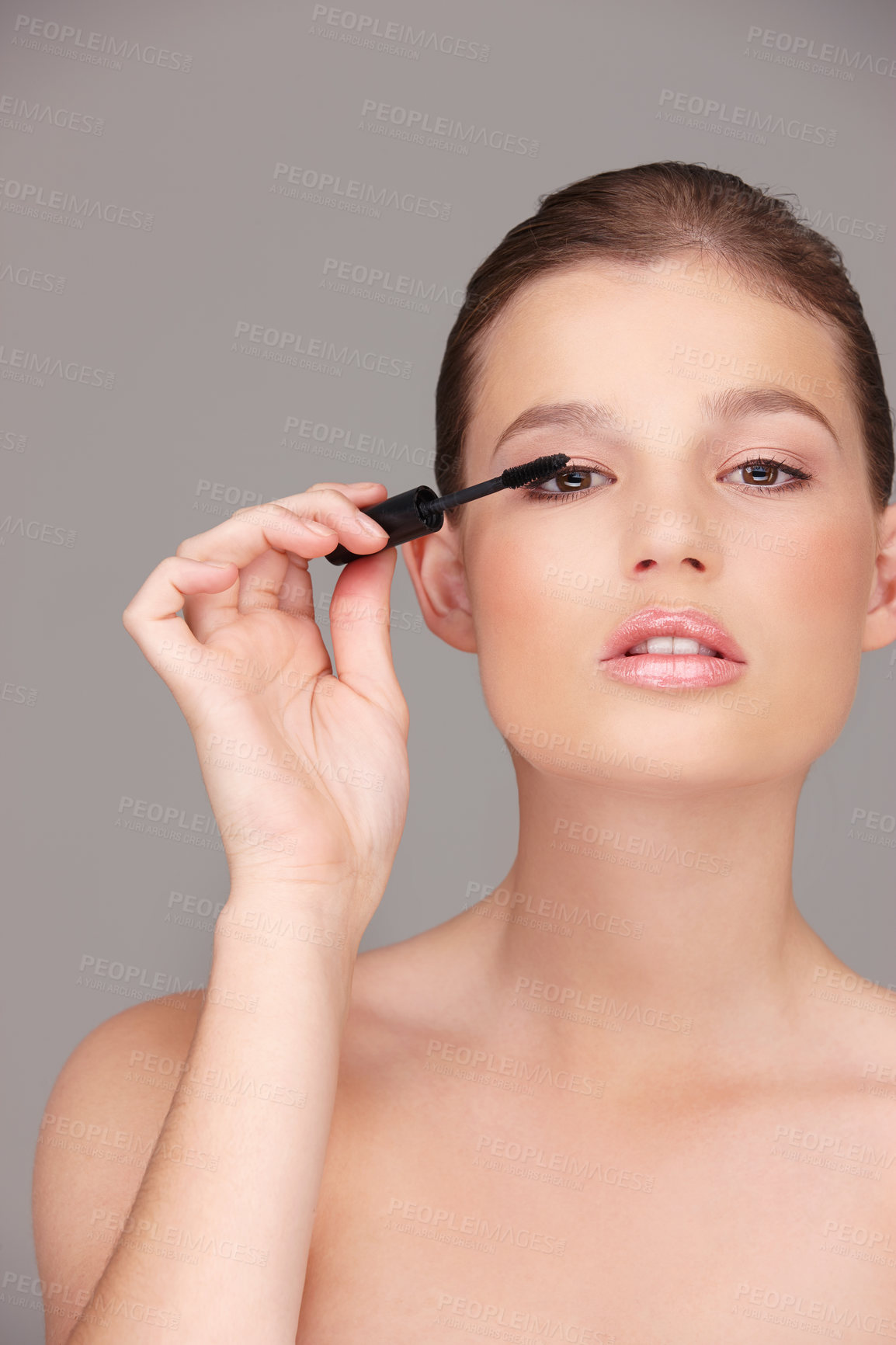 Buy stock photo Studio portrait of a woman using mascara isolated against a grey background. Young beautiful model applying makeup before a cosmetic shoot. Caucasian female beauty influencer doing a style tutorial