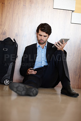 Buy stock photo A young businessman sitting on the floor and using his mobile while holding his tablet
