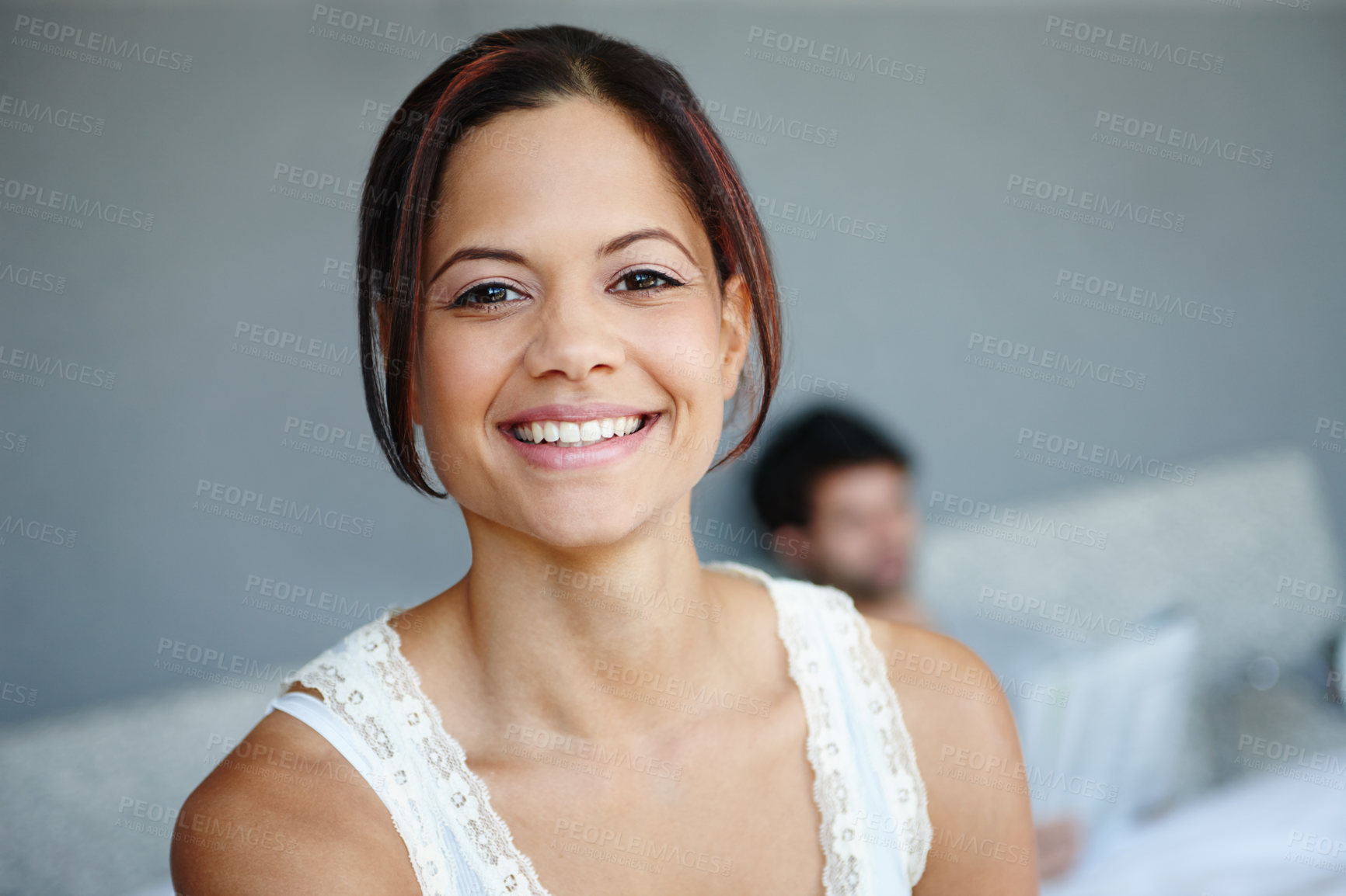 Buy stock photo Portrait of a smiling woman on the side of her bed with her boyfriend in the background