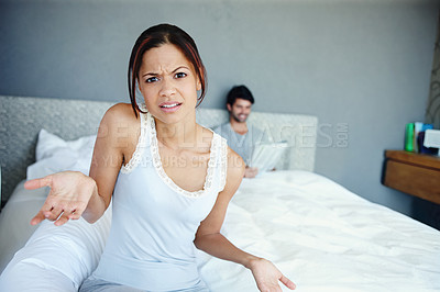 Buy stock photo Portrait of a confused woman sitting on the side of her bed with her boyfriend in the background