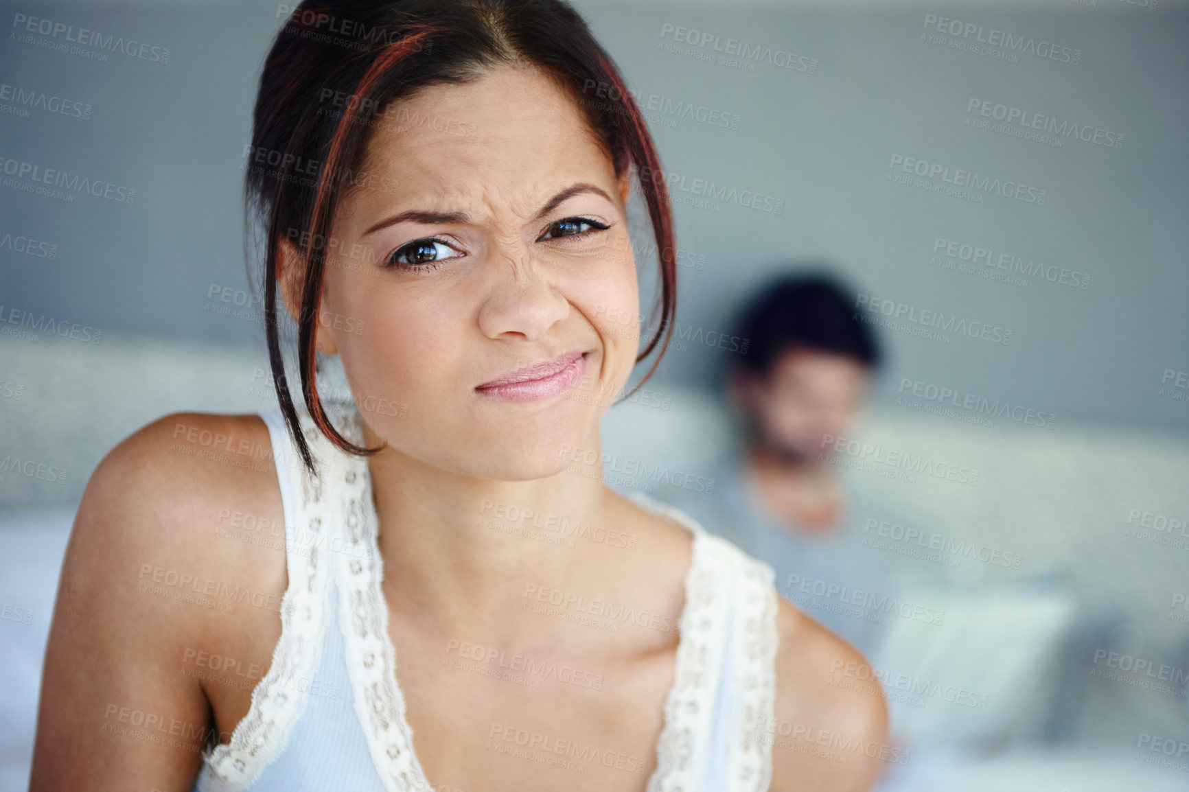 Buy stock photo Portrait of a woman scrunching up her face sitting in her bedroom with her boyfriend in the background