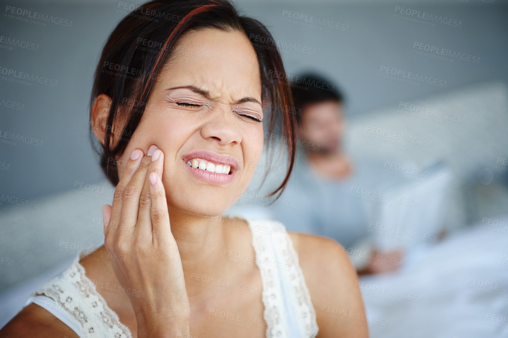 Buy stock photo Shot of a woman sitting on the side of her bed with bad toothache with her boyfriend in the background