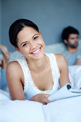 Buy stock photo Portrait of a smiling woman lying on her bed with a magazine in front of her with her boyfriend in the background