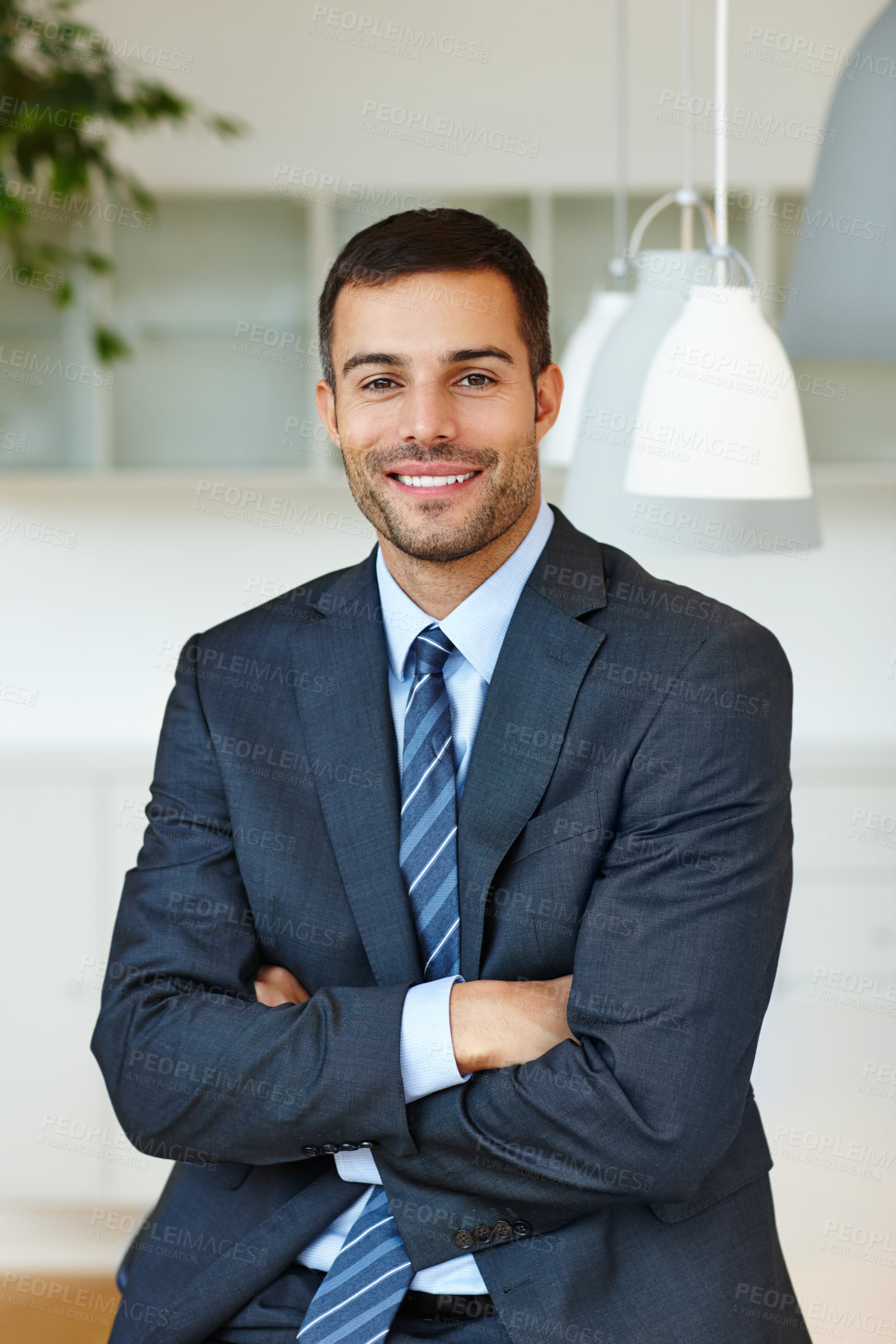 Buy stock photo Portrait of a proud, handsome young businessman with his arms crossed in an office at work. Confident male business professional wearing a suit and smiling while at his workplace. Proud boss at work