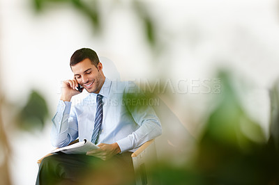 Buy stock photo A smiling young businessman on his mobile while sitting and reading a magazine