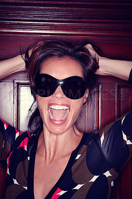 Buy stock photo Frustrated model screaming and pulling her hair while feeling angry at a party. One fashionable woman wearing sunglasses and shouting with her mouth open while standing against a wooden door inside. 