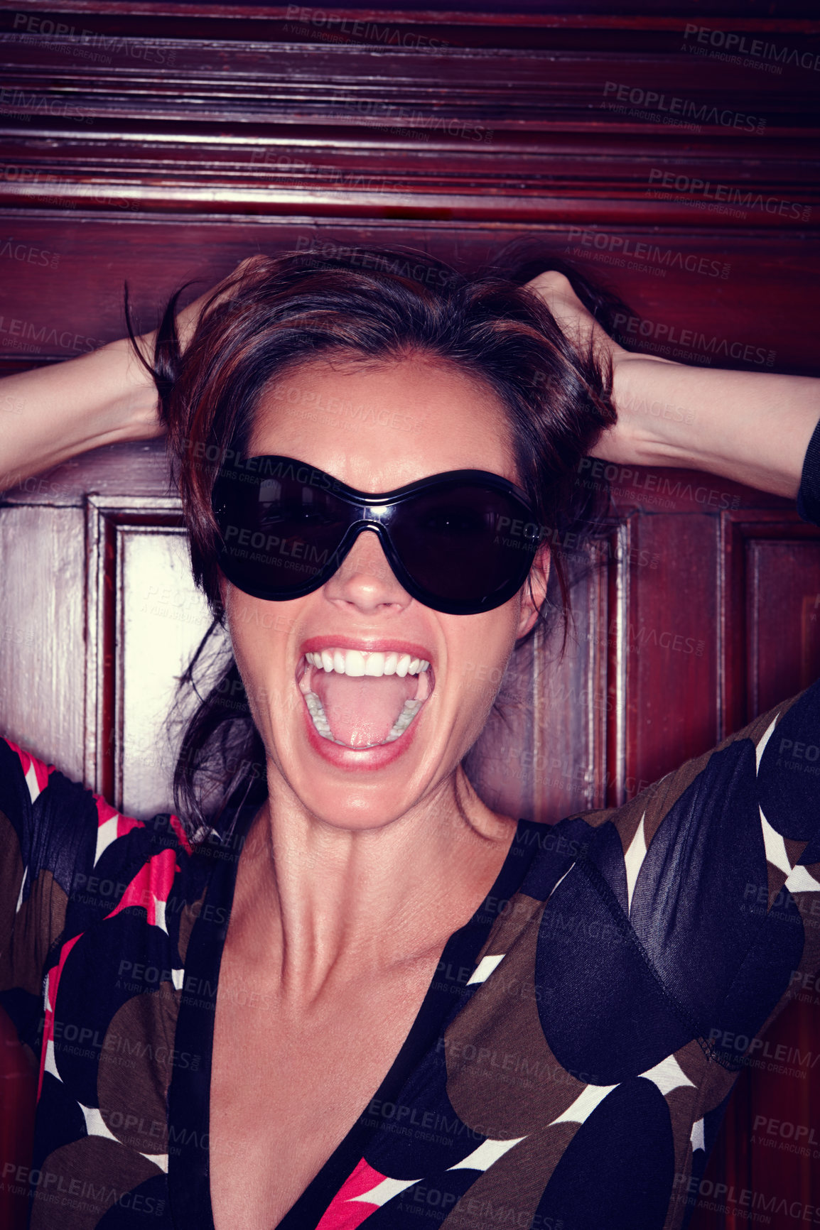 Buy stock photo Frustrated model screaming and pulling her hair while feeling angry at a party. One fashionable woman wearing sunglasses and shouting with her mouth open while standing against a wooden door inside. 