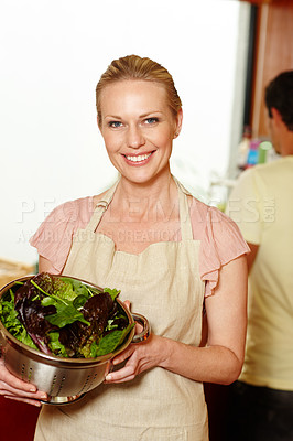 Buy stock photo A beautiful woman standing in her kitchen holding a bowl of lettuce while her husband stands in the background