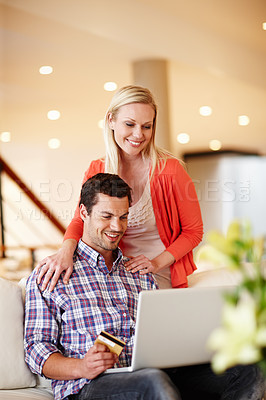 Buy stock photo A handsome man sitting on the couch with his laptop holding a credit card while his wife stands behind him