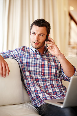 Buy stock photo A handsome man speaking on his cellphone while sitting with his laptop on the couch