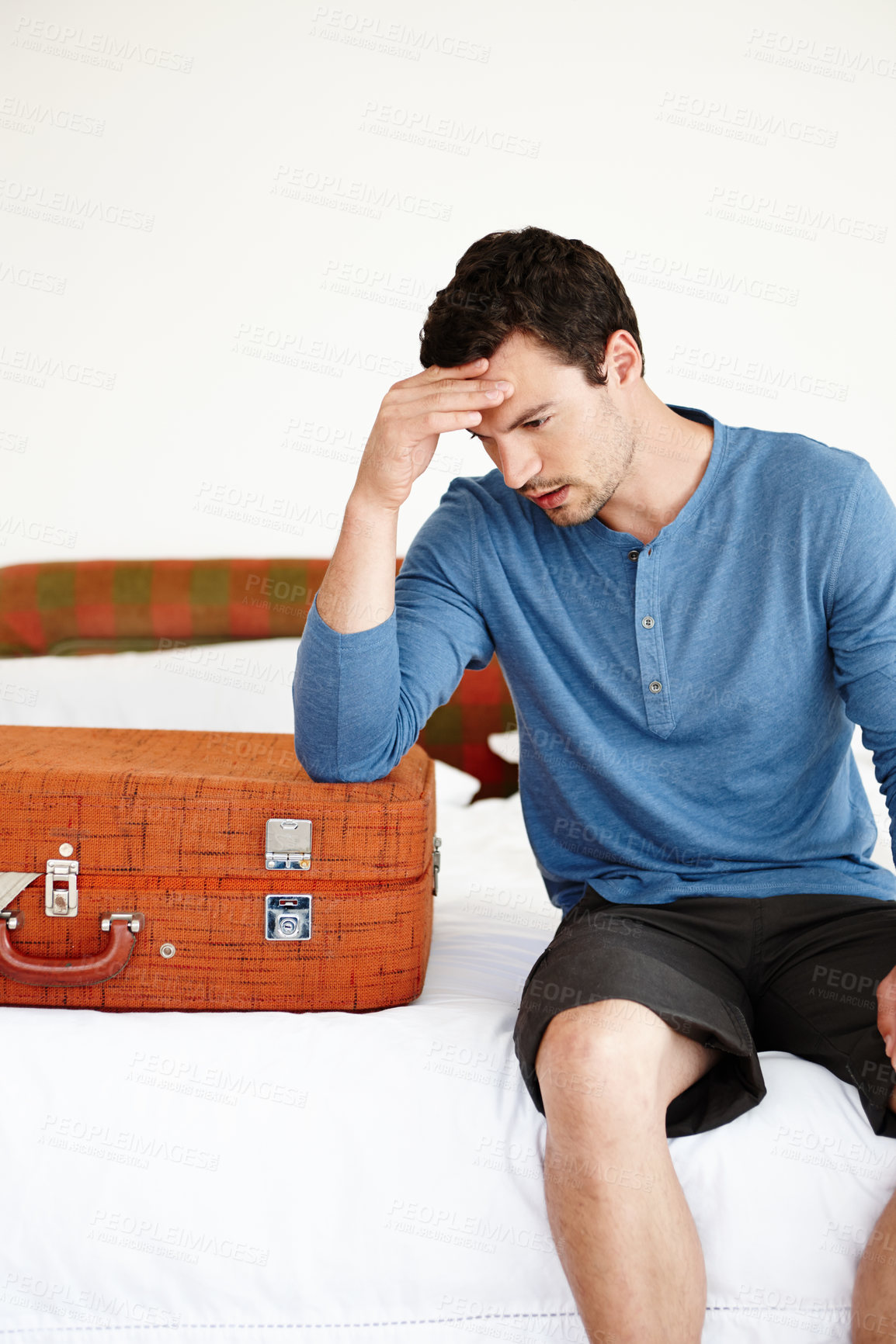 Buy stock photo A pensive young man sitting on his bed alongside a suitcase