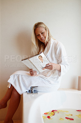 Buy stock photo A beautiful young woman reading a magazine while she sits on the edge of a luxurious bath filled with petals