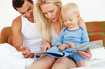 Buy stock photo Two young parents sitting on the bed reading a story book to their young son