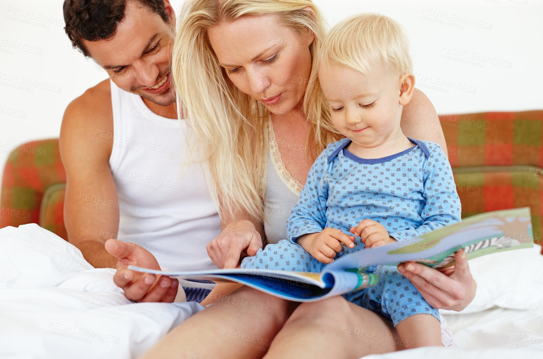 Buy stock photo Two young parents sitting on the bed reading a story book to their young son