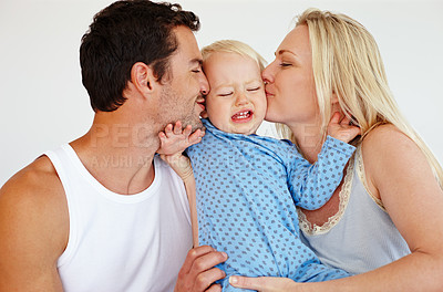 Buy stock photo Two loving parents kissing their son on his cheeks while he looks upset and tries to push them away