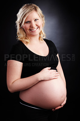 Buy stock photo Portrait of a pregnant woman holding her stomach isolated with a black background. Happy and smiling female expecting a baby standing alone inside a room. Attractive mom with an unborn child