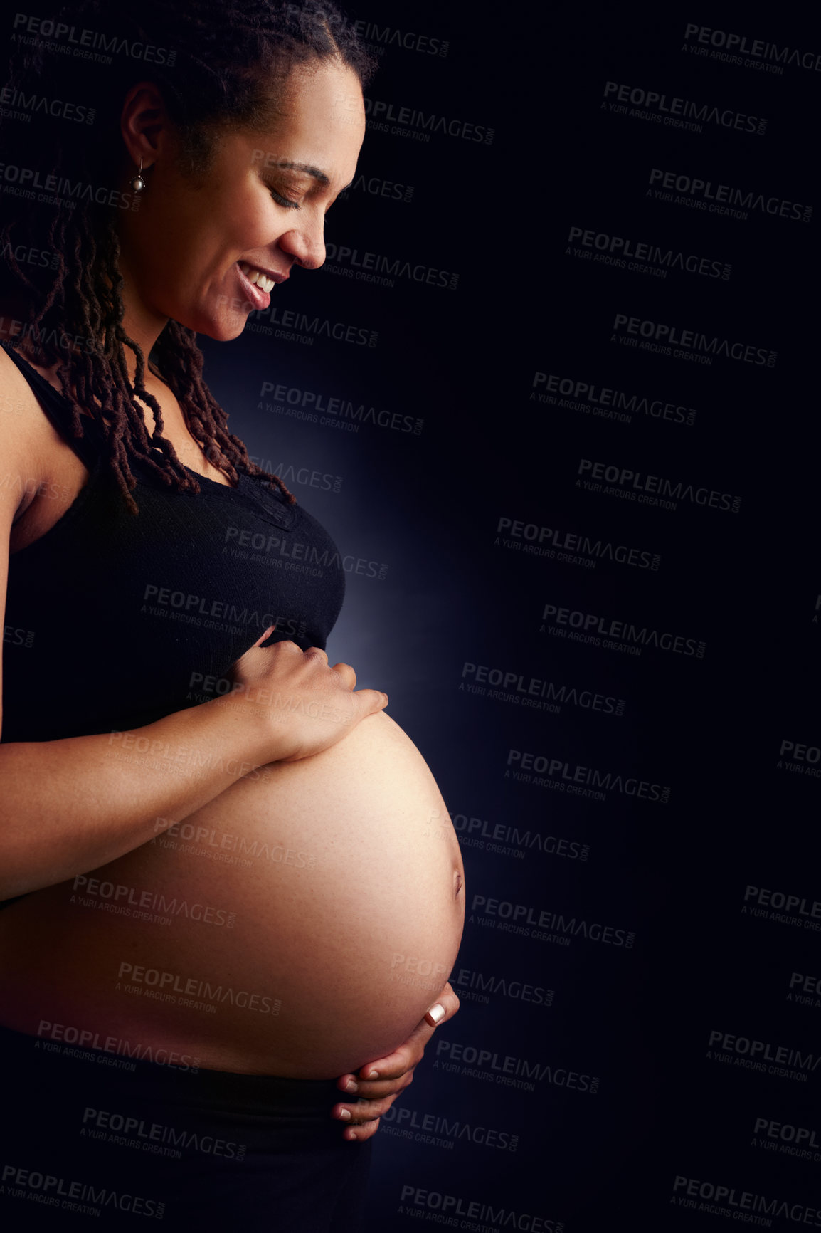 Buy stock photo A side profile of a happy pregnant woman holding her bare belly and looking down. Woman expecting a baby isolated with a black studio background. Lady with an unborn child standing alone in a room