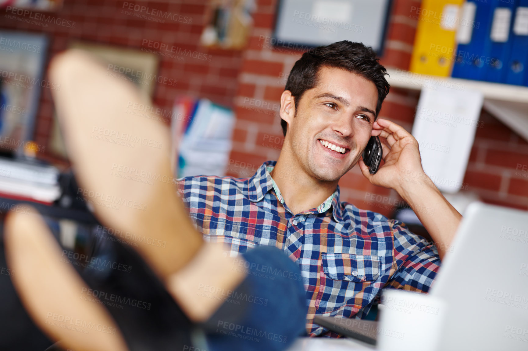 Buy stock photo A handsome young designer taking a call from a client while sitting at his desk with his feet up