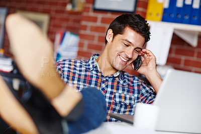 Buy stock photo A handsome young designer taking a call from a client while sitting at his desk with his feet up